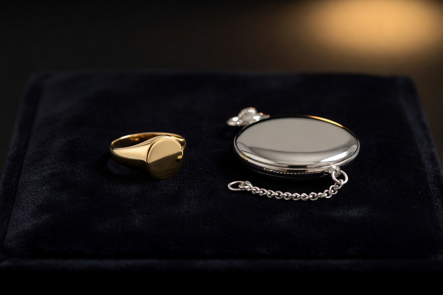 Polished plain gold signet ring beside a closed antique silver pocket watch with smooth cover and short fob chain on dark navy velvet jewelry display surface lit by a single warm amber spotlight from above