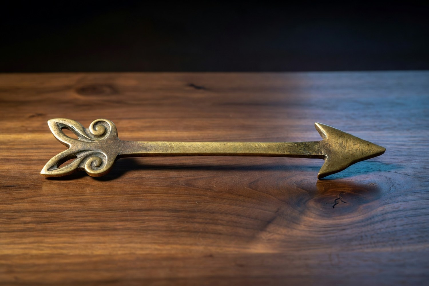 Antique hand forged brass directional arrow ornament lying horizontally on a polished walnut wooden surface with the arrow pointing right showing a forged arrowhead and decorative scrollwork at the base with aged patina lit by soft warm directional light from the upper left