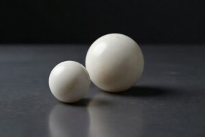 Two pure pale cream marble spheres of different sizes resting alone on a smooth polished dark charcoal slate surface with the smaller sphere in the left foreground and the larger sphere behind and to the right under soft directional cool light from the upper left casting subtle long cool gray shadows across the slate