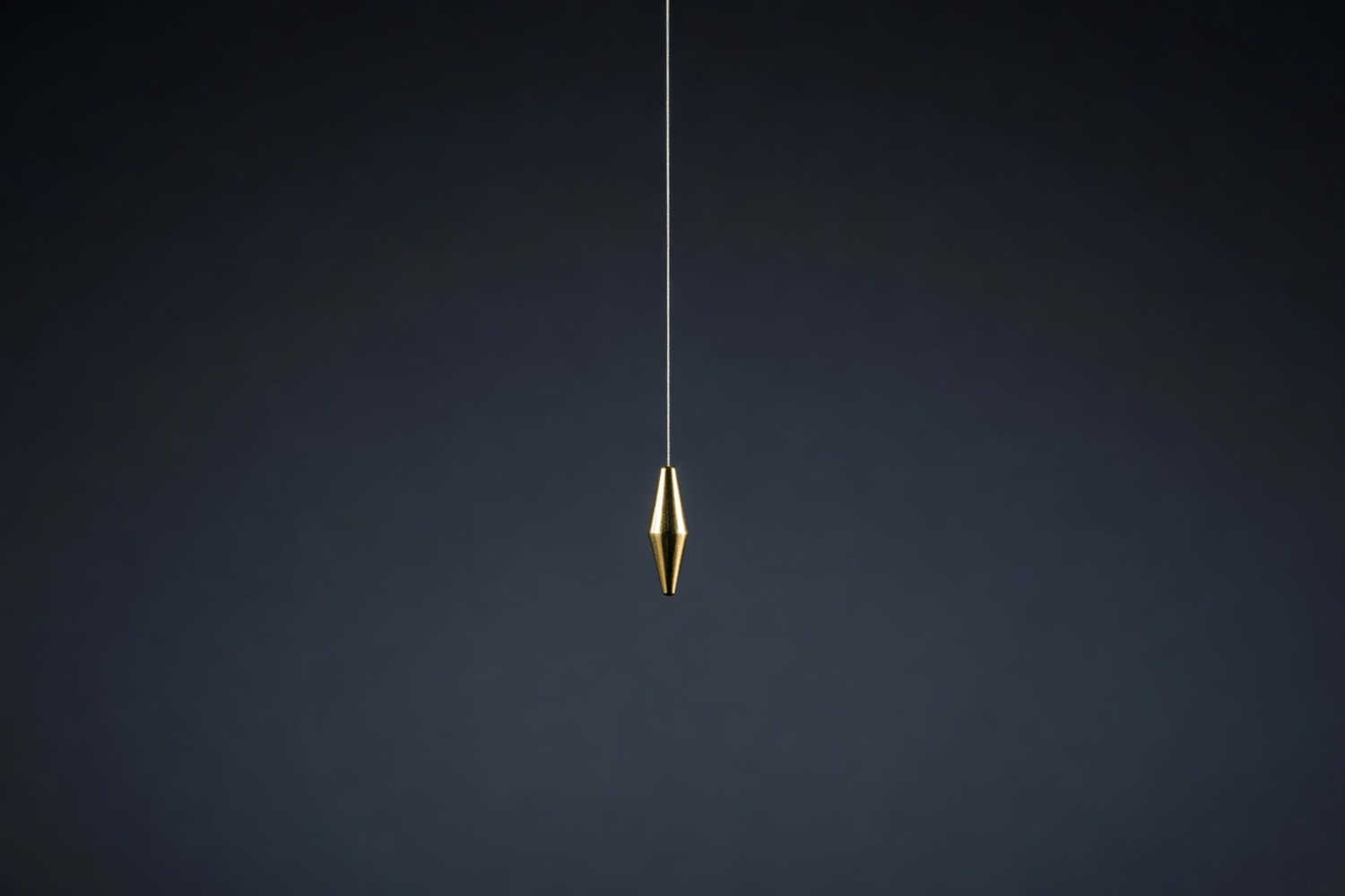 Single polished brass plumb bob in a faceted teardrop shape suspended from a fine silver thread descending from the upper edge of the frame against a deep charcoal gradient background with soft cool directional light from the upper left catching warm specular highlights on the brass