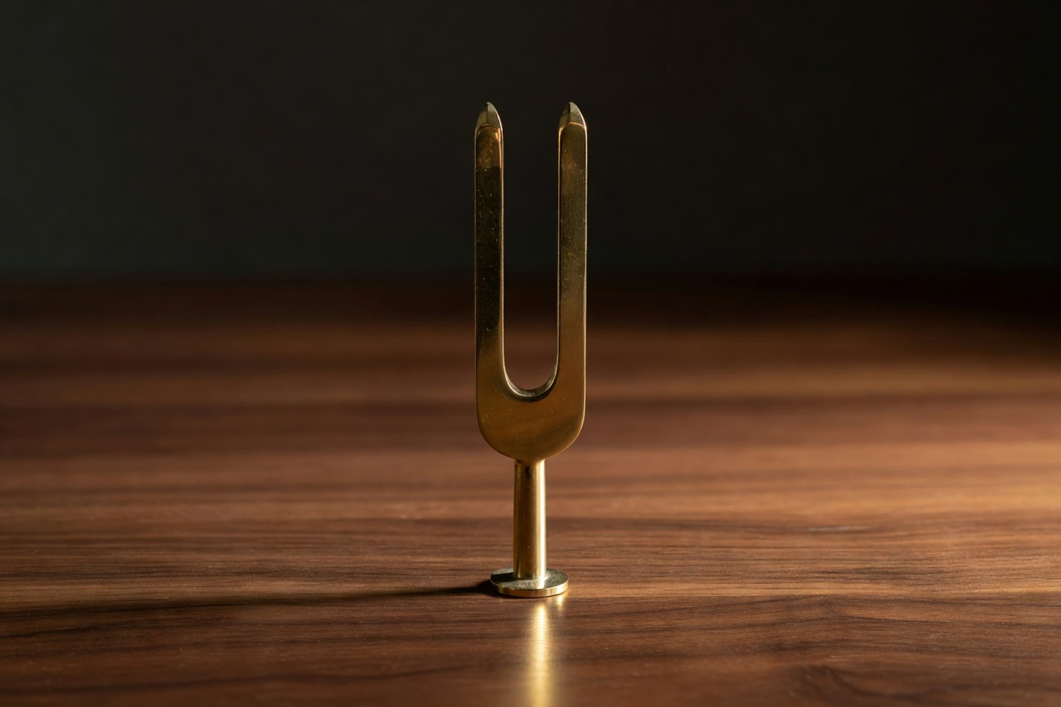 Single polished brass tuning fork standing upright alone on a smooth polished walnut wooden surface with classic two tine U shape rising from a slim cylindrical stem on a small round base under warm directional light from the right with brilliant warm specular highlights on the brass and a gentle warm shadow stretching to the left across the wood