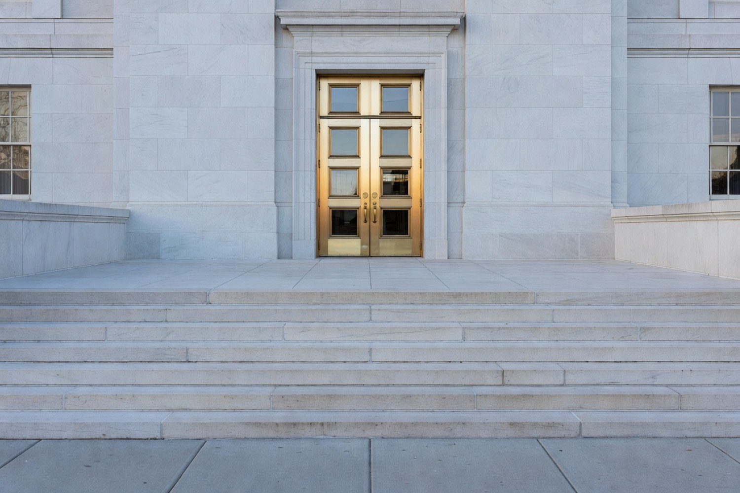 Front entrance approach to a major U.S. government office building photographed from outside on the public sidewalk looking toward a set of tall classical brass framed glass doors at the top of a short wide stone staircase with white limestone facade plain classical rectangular window openings on either side under cool overcast late afternoon daylight