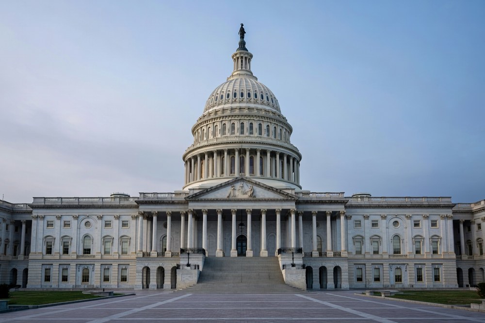 United States Capitol Building exterior in Washington DC photographed from a low three quarter angle on the central staircase approach in cool overcast late afternoon light with the iconic white marble dome dominating the upper portion of the frame the central pedimented portico and Corinthian columns visible at center and the broad central staircase descending in the foreground under a cool slightly hazy sky