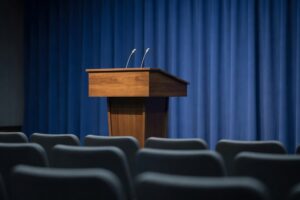 Empty press briefing room set up for an imminent press conference photographed from the back of the room looking forward toward a polished walnut speaking podium positioned in the center with two slim upright broadcast microphones angled toward the camera against a deep cobalt blue draped fabric backdrop wall with rows of empty journalist chair backs in soft foreground bokeh under soft warm overhead studio lighting