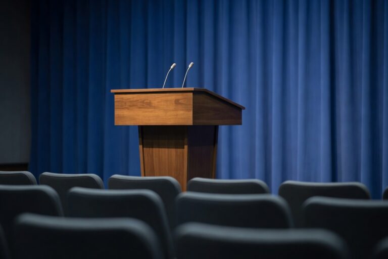 Empty press briefing room set up for an imminent press conference photographed from the back of the room looking forward toward a polished walnut speaking podium positioned in the center with two slim upright broadcast microphones angled toward the camera against a deep cobalt blue draped fabric backdrop wall with rows of empty journalist chair backs in soft foreground bokeh under soft warm overhead studio lighting