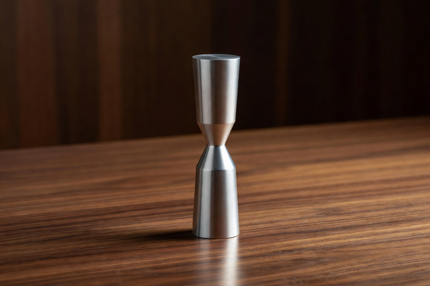 Single brushed aluminum abstract sculptural form shaped like a tall slim hourglass body with two stacked cone shapes meeting at a narrow waist standing upright on a polished walnut wooden surface under warm directional light from the upper right catching the polished surface with brilliant cool specular highlights