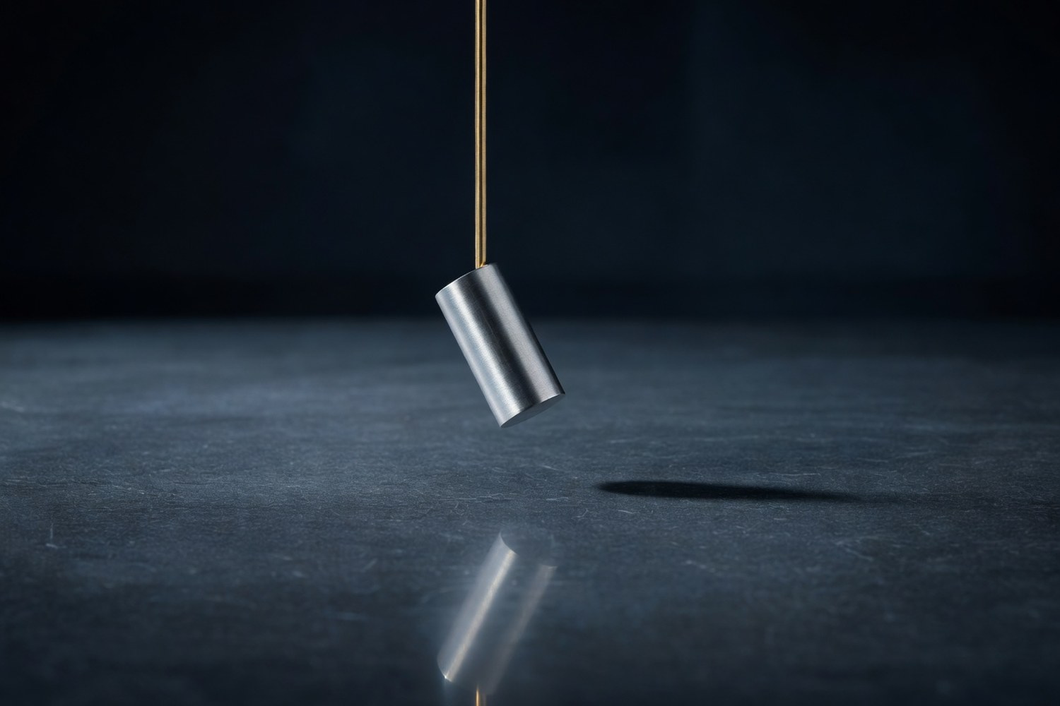 Single brushed aluminum cylindrical pendulum bob suspended from a slim brass pivot rod above a polished dark slate surface captured mid swing with the bob slightly off center suggesting a moment frozen in motion under soft cool directional light from the upper left with a subtle reflection on the slate and a soft shadow stretching to the right
