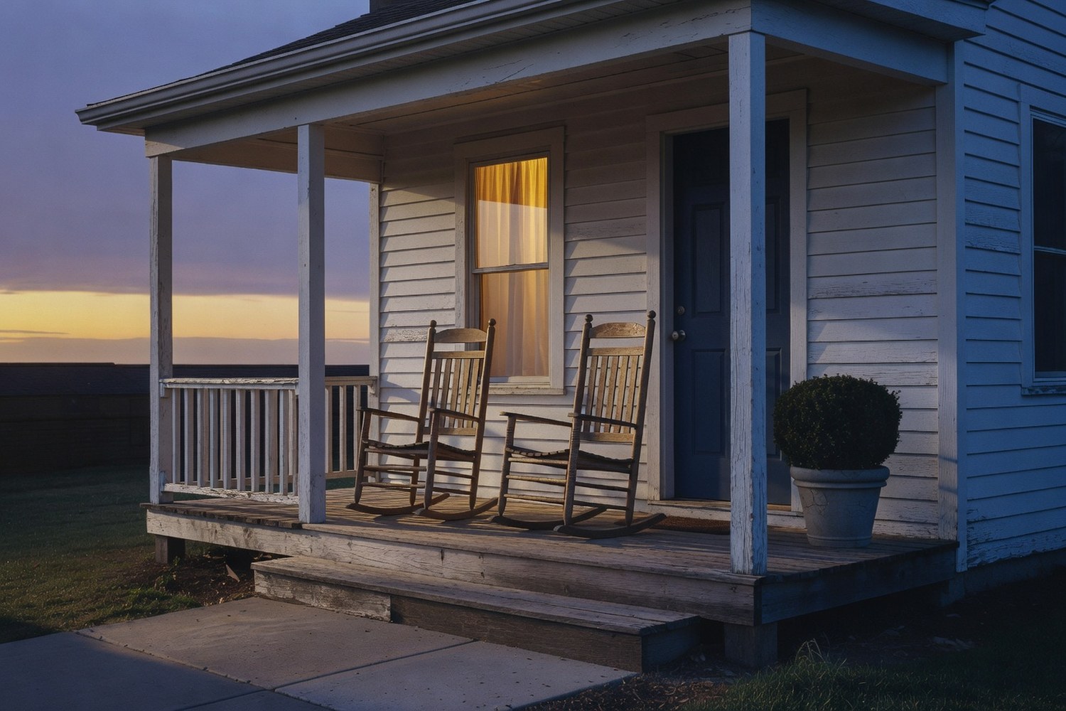 Modest American residential front porch at dusk with weathered white clapboard siding navy blue door warm amber window glow and two empty wooden rocking chairs against a blue-violet twilight sky