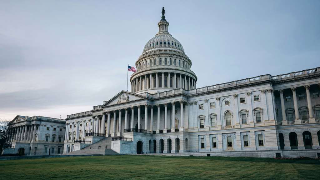 Wide editorial photograph of the United States Capitol Senate side photographed from low on the east lawn looking up at the Senate wing dome and pediment under cool overcast late afternoon daylight white marble columns clearly visible flag flying institutional formality no people present
