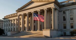 Wide exterior view of the United States Treasury Department building with its neoclassical limestone columns and a large American flag, photographed in dramatic golden late afternoon light with long shadows across the facade