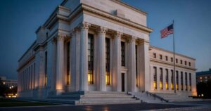 The marble facade of the Federal Reserve Eccles Building in Washington photographed at dusk, neoclassical Corinthian columns lit by warm interior light beneath a deep blue evening sky with an American flag on a pole to the right and a broad staircase in the foreground