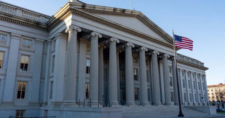 Wide exterior photograph of the United States Treasury Department building in Washington DC, neoclassical stone columns and facade lit by cool late afternoon light with an American flag at the side, photographed in a documentary financial-news style under a muted slate-blue sky