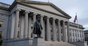 The United States Treasury Department building in Washington DC photographed wide in cool overcast daylight, its neoclassical granite facade and tall columns visible with the Alexander Hamilton statue and an American flag in front under a pale gray sky