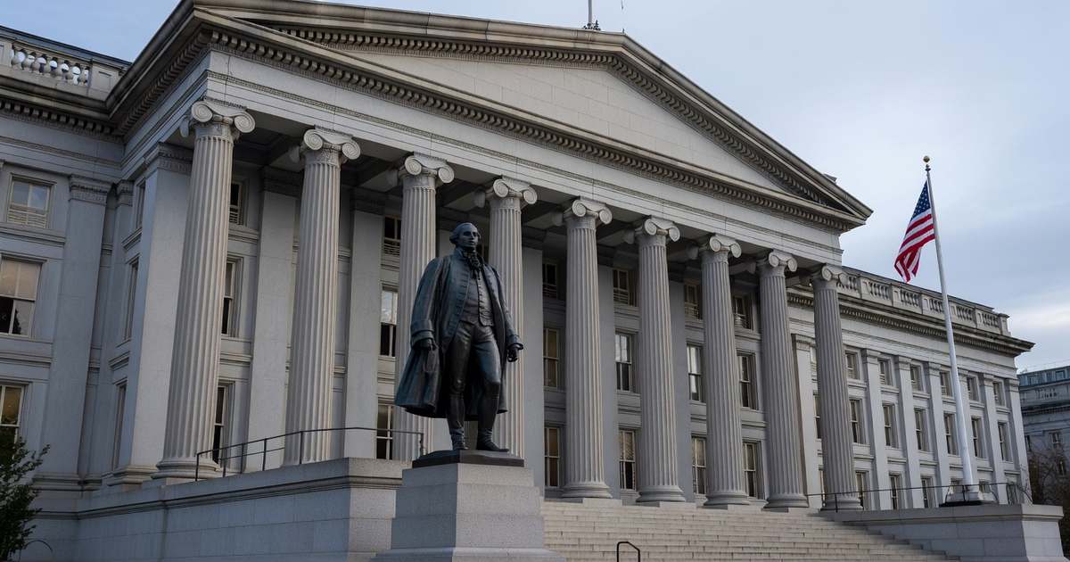 The United States Treasury Department building in Washington DC photographed wide in cool overcast daylight, its neoclassical granite facade and tall columns visible with the Alexander Hamilton statue and an American flag in front under a pale gray sky