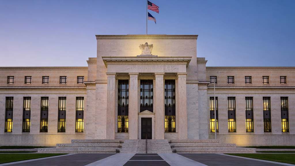 The neoclassical white marble facade of the United States Federal Reserve Eccles Building in Washington photographed head-on at blue hour with tall fluted columns, warmly lit interior windows, and an American flag flying above the central pediment