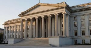 The U.S. Treasury Department building in Washington at sunrise, its neoclassical stone columns lit by golden morning light.