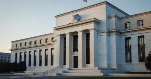 The white marble neoclassical facade of the Federal Reserve Marriner S. Eccles Building in Washington under soft morning light with columns and an American flag