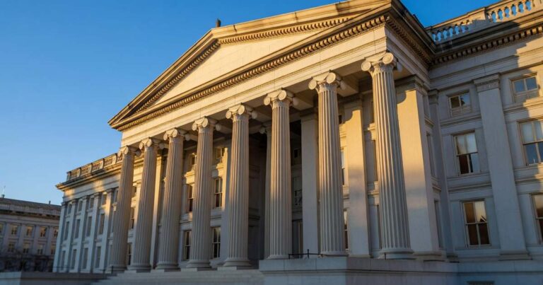 The neoclassical facade of the United States Treasury Department building photographed from a low angle in warm early morning sunlight, its tall stone columns casting long shadows against a clear blue sky.