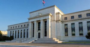 Wide photograph of the Federal Reserve headquarters neoclassical marble building in Washington with tall granite columns and an American flag under clear morning light