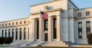 The marble neoclassical facade of the United States Federal Reserve headquarters building in Washington photographed from the plaza with tall fluted stone columns, an American flag on a pole, wide granite steps, and a clear sky in warm late afternoon light