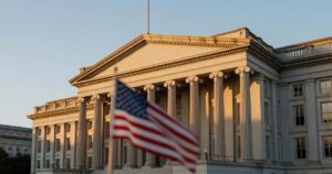 The United States Treasury building neoclassical stone facade with tall columns in Washington photographed at golden hour with a softly blurred American flag in the foreground under a clear sky