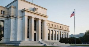 Wide morning view of a neoclassical United States Federal Reserve headquarters building with a white marble facade, tall columns, and an American flag on a pole to the right under a clear pale sky