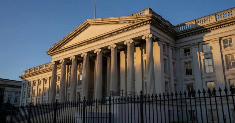 Wide golden-hour view of the neoclassical United States Treasury Department building in Washington DC, its tall stone columns and portico lit by warm low afternoon sunlight against a deep blue sky, with an iron perimeter fence blurred in the foreground.