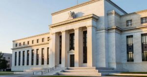 The white marble neoclassical Federal Reserve headquarters building in Washington DC photographed at sunrise, with tall columns, bronze doors and a wide stone staircase lit by low golden morning light against a pale blue sky