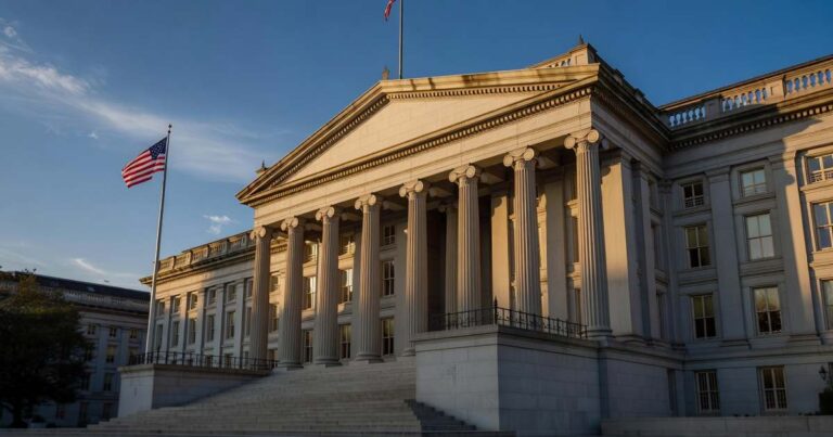 The neoclassical United States Treasury Building in Washington lit by low golden hour sunlight, its tall stone columns and pediment in warm relief against a deep blue late summer sky, with an American flag catching the light beside the entrance steps.