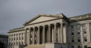 The columned stone facade of the United States Treasury building in late afternoon light beneath an overcast gray sky