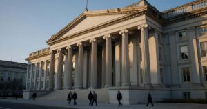 Late afternoon sunlight rakes across the limestone columns of the United States Treasury Building in Washington as pedestrians pass on the sidewalk below.
