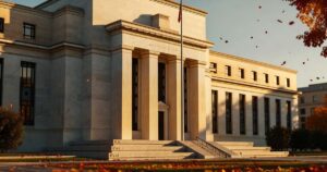 Late afternoon sunlight rakes across the limestone columns and stepped entrance of the Federal Reserve Board headquarters in Washington, with autumn leaves scattered on the lawn and a bare flagpole rising in front of the portico.