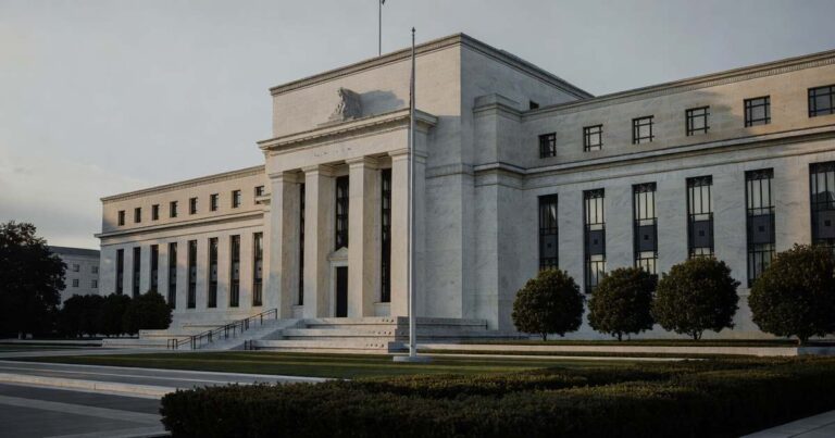 The neoclassical limestone facade of the Federal Reserve Marriner S. Eccles Building in Washington photographed at early morning with tall fluted columns deep window recesses raking dawn light across the stone long cool shadows falling on the empty plaza a bare flagpole and low trimmed hedges in the foreground beneath a pale overcast sky