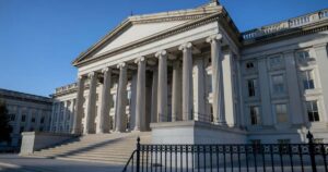The neoclassical limestone west facade of the United States Treasury building in Washington DC photographed from a low angle in early morning light, tall fluted Ionic columns above wide stone steps, a black wrought iron fence in the foreground and clear blue sky overhead.