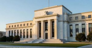 The neoclassical marble facade of the Federal Reserve headquarters in Washington DC lit by low golden sunrise light, tall columns casting long shadows across the stone steps under a clear pale blue sky.