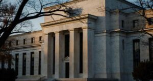 The neoclassical marble facade of the Federal Reserve Board headquarters in Washington DC photographed in late afternoon autumn light, with bare tree branches in the foreground and deep shadows falling between the building's tall columns.