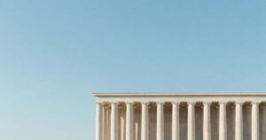Upper portion of a classical United States federal government building photographed from below with a row of smooth pale limestone columns standing against a wide clear gradient blue morning sky that fills the upper two thirds of the frame under soft even directional sunlight