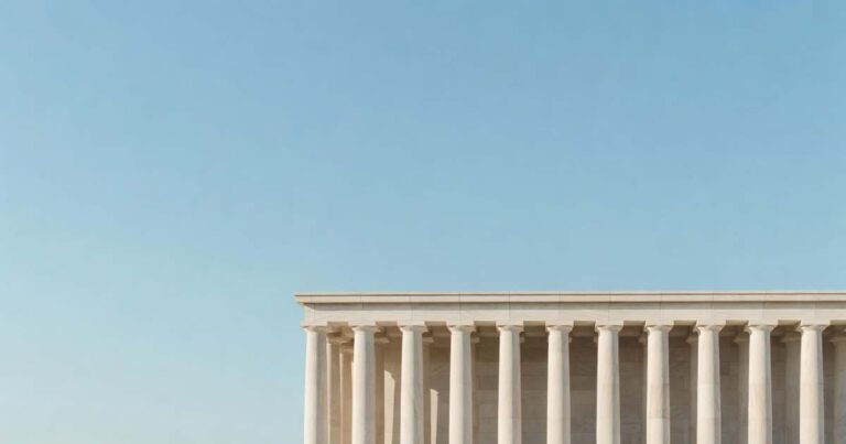 Upper portion of a classical United States federal government building photographed from below with a row of smooth pale limestone columns standing against a wide clear gradient blue morning sky that fills the upper two thirds of the frame under soft even directional sunlight