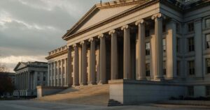 The United States Treasury Department building in Washington D.C. photographed in wide composition at golden hour with tall neoclassical stone columns along the facade, long angled shadows falling across the limestone steps, and warm late afternoon light breaking through an overcast autumn sky above the roofline