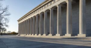 Wide view of a large neoclassical United States federal government building photographed from across an empty paved plaza in early morning light, a long colonnade of tall fluted stone columns receding into the distance and casting deep diagonal shadows across the pavement, bare tree branches entering at the left edge against a clear pale blue sky