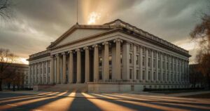 Wide exterior view of the neoclassical United States Treasury Department building in Washington photographed from across the street in warm late afternoon October light with tall stone columns casting long shadows down the facade, bare autumn tree branches entering the frame at one edge, and an overcast sky broken by a band of gold light above the roofline