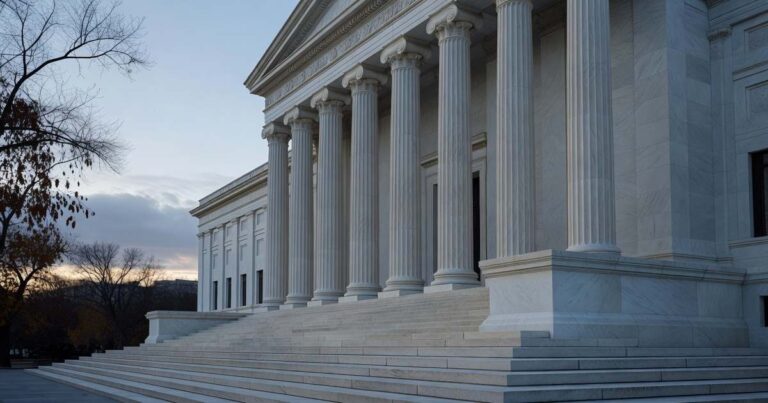 Neoclassical white marble facade of a United States central bank headquarters building photographed at early morning from the base of its wide granite steps looking up along a row of tall fluted stone columns supporting a raised portico with bare autumn tree branches framing the left edge under cool blue grey overcast dawn light