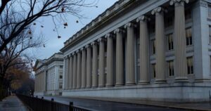 Wide exterior view of the neoclassical United States Treasury building in Washington DC photographed from across the street on an overcast late autumn morning with tall fluted stone columns running the length of the colonnade, bare maple branches framing the left edge, wet dark pavement in the foreground and a muted grey blue sky overhead
