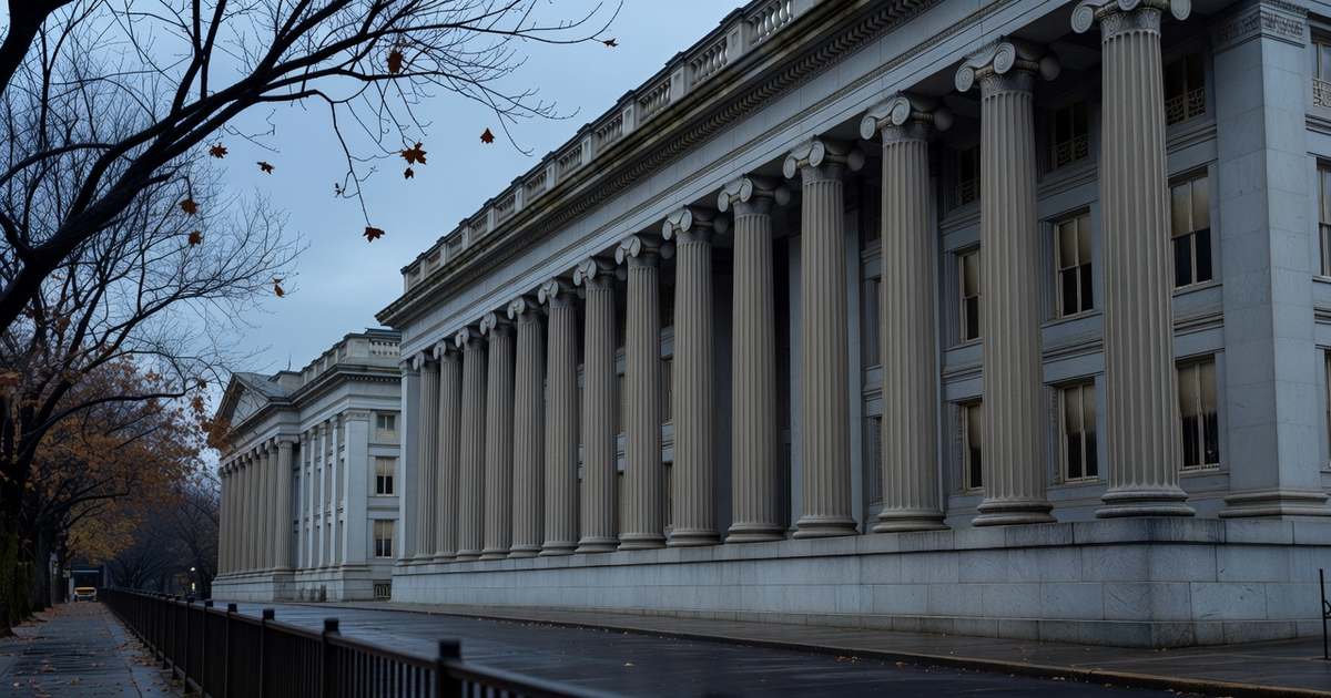 Wide exterior view of the neoclassical United States Treasury building in Washington DC photographed from across the street on an overcast late autumn morning with tall fluted stone columns running the length of the colonnade, bare maple branches framing the left edge, wet dark pavement in the foreground and a muted grey blue sky overhead