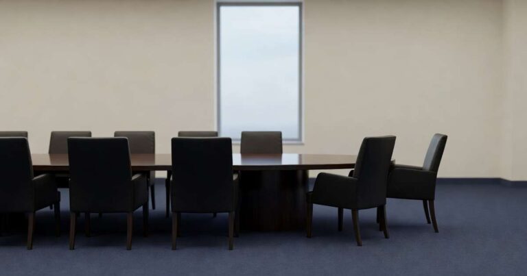 Empty formal government boardroom with a long polished dark wood oval conference table surrounded by high backed black leather chairs, two chairs at the right pushed back and angled away from the table while the rest sit squarely, a tall plain window at the far wall casting flat overcast afternoon light across cream walls and navy carpet